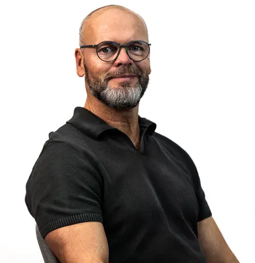 Portrait of a bald man with a gray beard and glasses, wearing a black polo shirt, looking at the camera with a slight smile.